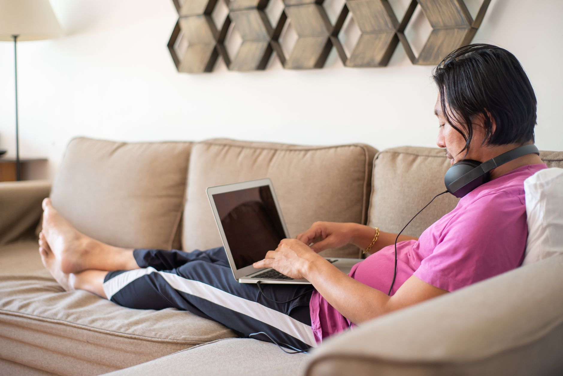An Asian man in a pink shirt working on his laptop while wearing headphones, relaxing on a couch indoors.