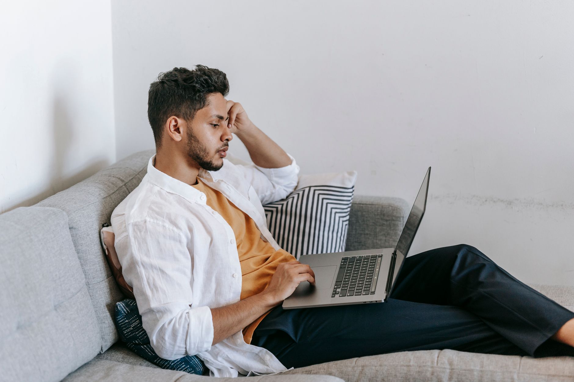 Side view of a serious young Hispanic male remote employee sitting on a sofa and using a laptop while working distantly from home.
